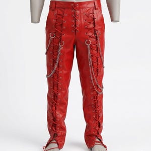 May include: Bright red leather trousers featuring a lace-up design on the front and sides. Silver chains hang from the waist and sides. The trousers have a fitted shape and are displayed on a mannequin.