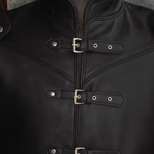 以下が含まれることがあります： ハイカラーと装飾的なステッチが施された黒い革のジャケットのクローズアップ。 ジャケットには、安全なフィット感のための3つのシルバーバックルとアイレットが付いています。 デザインは詳細です。