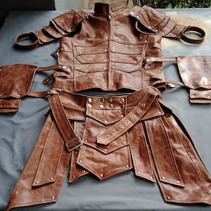 K&ouml;nnte beinhalten: Ein braunes Lederkost&uuml;m-Set, bestehend aus einer Jacke mit mehreren Taschen und Schulterpanzern, einem G&uuml;rtel und einem rockartigen Unterteil. Das Set ist mit Nieten und Schnallen versehen, was auf ein mittelalterliches oder Fantasy-Thema hindeutet.