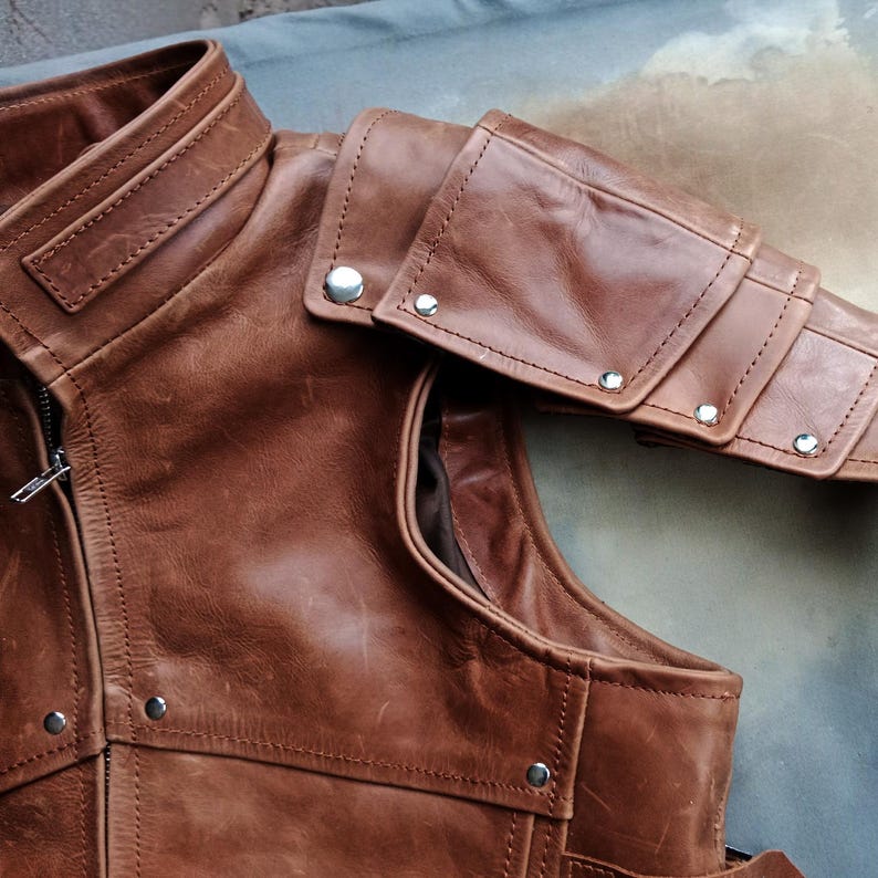 K&ouml;nnte beinhalten: Nahaufnahme einer braunen Lederweste mit hohem Kragen und Schulterpanzerung. Die Weste hat einen Rei&szlig;verschluss und ist mit silbernen Nieten versehen. Das Leder hat einen Used-Look, der ihm ein Vintage-Aussehen verleiht.