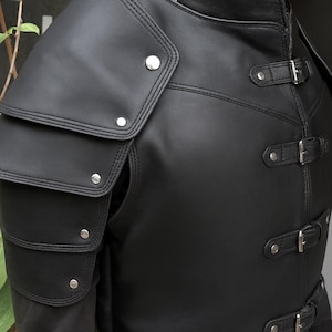 以下が含まれることがあります： 黒い革のジャケット。肩には重ねられたアーマー、銀色の金属リベットが付いています。ジャケットはハイカラーで、バックル付きの調節可能なストラップが付いています。デザインは中世の鎧を連想させます。