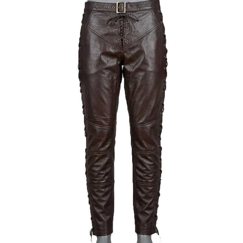 May include: Brown leather pants with a lace-up front and sides. The pants have a belt with a gold buckle. The leather appears to be distressed, giving the pants a vintage look. The pants are designed with multiple panels and stitching.