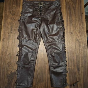 May include: Brown leather pants with a lace-up design on the sides and front. The pants have a fitted silhouette with a buckle closure at the waist. The leather has a slightly distressed appearance.