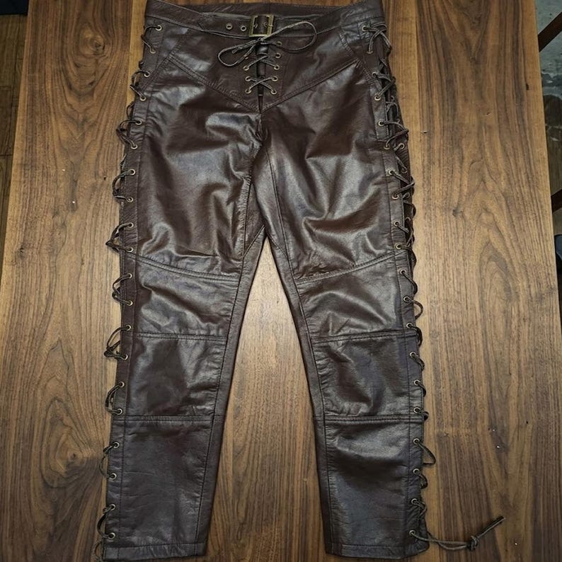 May include: Brown leather pants with a lace-up design on the sides and front. The pants have a fitted silhouette with a buckle closure at the waist. The leather has a slightly distressed appearance.