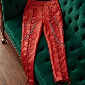 May include: Red leather trousers featuring a lace-up design and silver chain details. The trousers are displayed on a green velvet sofa, highlighting their distinctive and bold fashion statement.