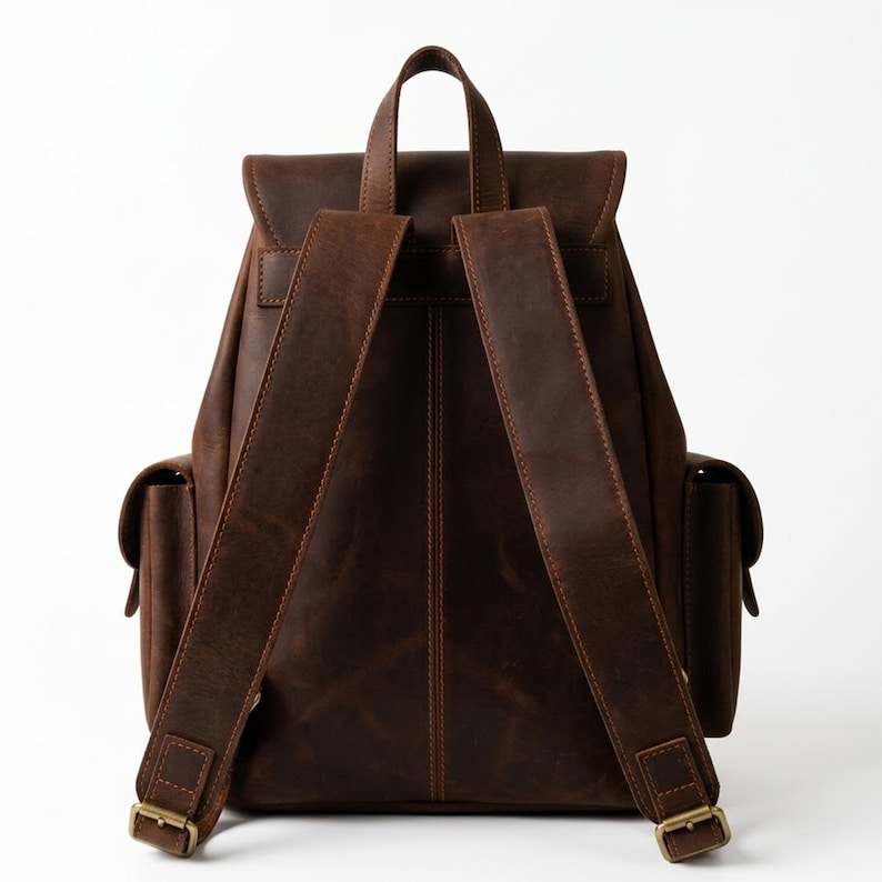 May include: A dark brown leather backpack featuring a top handle, two adjustable shoulder straps with metal buckles, and two side pockets. The backpack has a flap closure and visible stitching along the straps and the main body of the bag.