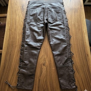 May include: Brown leather pants with lace-up detailing along the sides. The pants have two back pockets and a classic five-pocket design. The leather has a slightly distressed appearance.