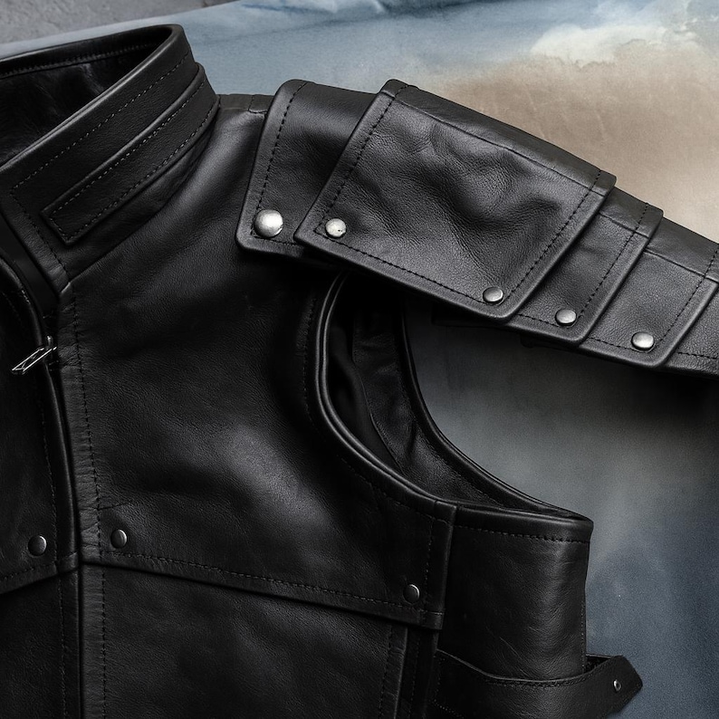 May include: Close-up of a black leather jacket with a high collar and shoulder armor detailing. The jacket features silver-toned snap closures and intricate stitching. The design suggests a vintage or biker-inspired aesthetic.
