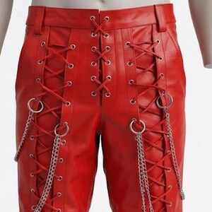 May include: Red leather trousers featuring a lace-up design. Silver chains and rings are attached to the lace-up details. The trousers have a classic five-pocket design and a belt loop.
