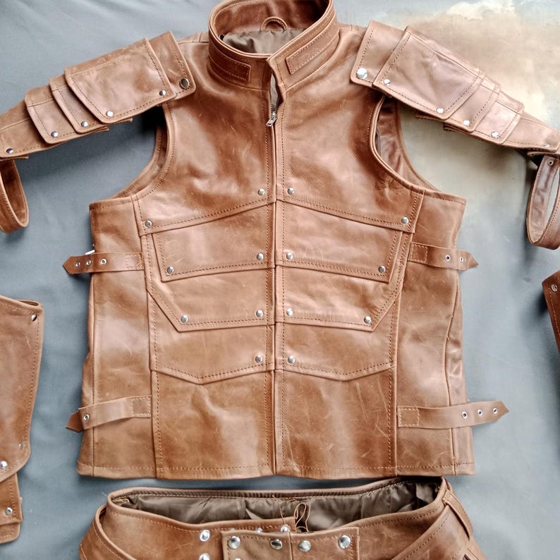 K&ouml;nnte beinhalten: Eine braune Lederweste mit hohem Kragen, Rei&szlig;verschluss und mehreren Taschen. Die Weste hat silberne Nietendetails und wird von passenden Schulterpanzern und einem G&uuml;rtel begleitet. Das Set ist f&uuml;r eine robuste, mittelalterliche &Auml;sthetik konzipiert.