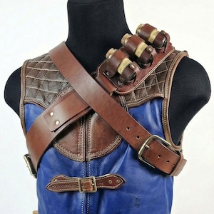 May include: A brown leather shoulder harness with three small cylindrical containers. The harness features a quilted pattern on the shoulder and a blue leather vest. A brown leather strap with a brass buckle crosses the chest.