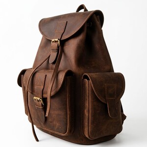 May include: A dark brown backpack featuring a fold-over flap with a buckle closure, two exterior side pockets with flap covers, and a central front pocket with a buckle. The bag is constructed with visible stitching and antique brass-toned hardware.