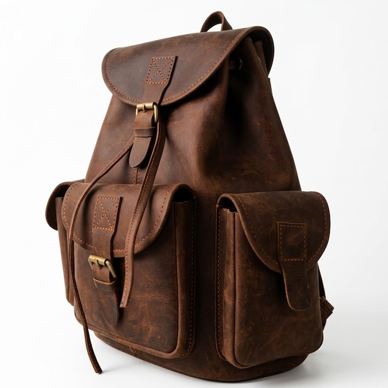 May include: A dark brown backpack featuring a fold-over flap with a buckle closure, two exterior side pockets with flap covers, and a central front pocket with a buckle. The bag is constructed with visible stitching and antique brass-toned hardware.