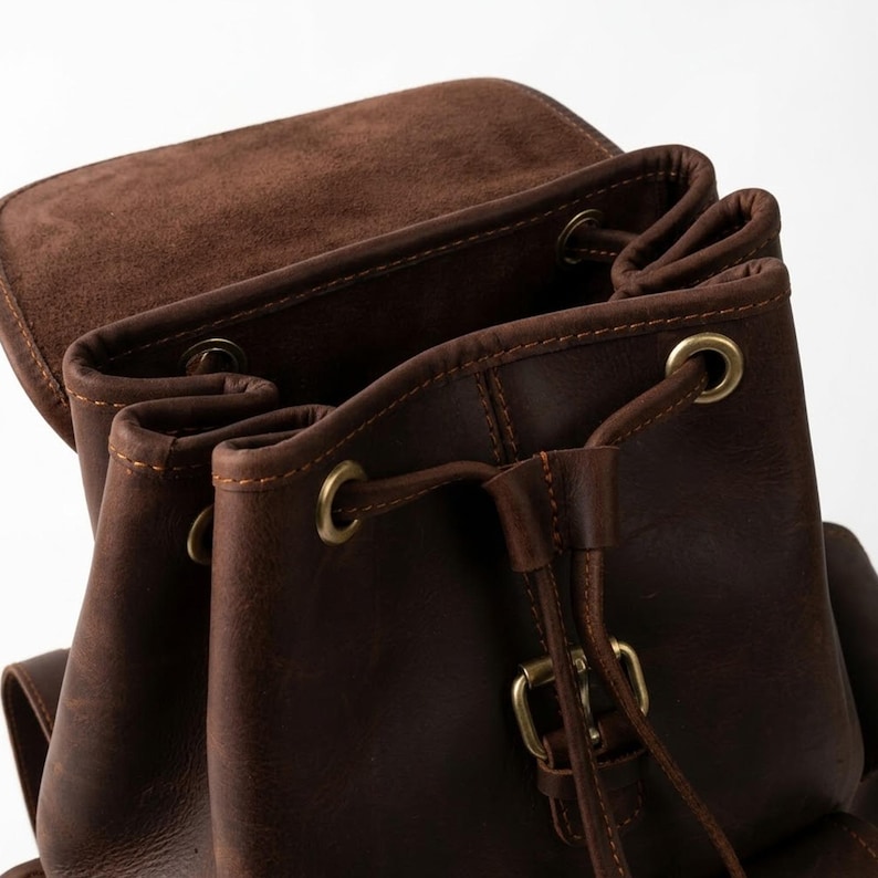 May include: A dark brown leather backpack featuring a fold-over flap, a drawstring closure with brass-toned metal eyelets, and a central vertical strap secured by a matching brass-toned metal buckle. The interior lining is visible.