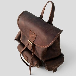 May include: A dark brown leather backpack featuring a large top flap with a buckle closure, a top handle, and two smaller exterior pockets on the front. The bag has visible stitching details and a matte, textured finish throughout the surface.