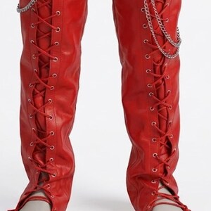 May include: Vibrant red knee-high boots featuring a lace-up design and silver chain detailing. The boots have a smooth, leather-like finish and a pointed toe. Red laces crisscross up the front of the boots.