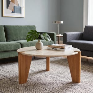 May include: A round coffee table with a light-colored stone top and three thick, angled wooden legs. On the table sits a small, light-colored ceramic vase containing a green monstera leaf and two stacked books titled Mid-Century Modern Design and Eames.