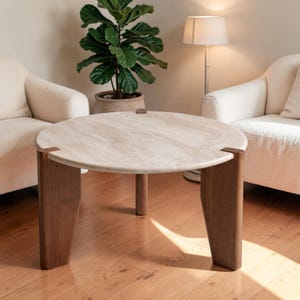 May include: A round coffee table featuring a beige, textured stone top with visible natural veining. The table is supported by three thick, dark brown wooden legs that connect to the stone surface with small, square metal brackets.
