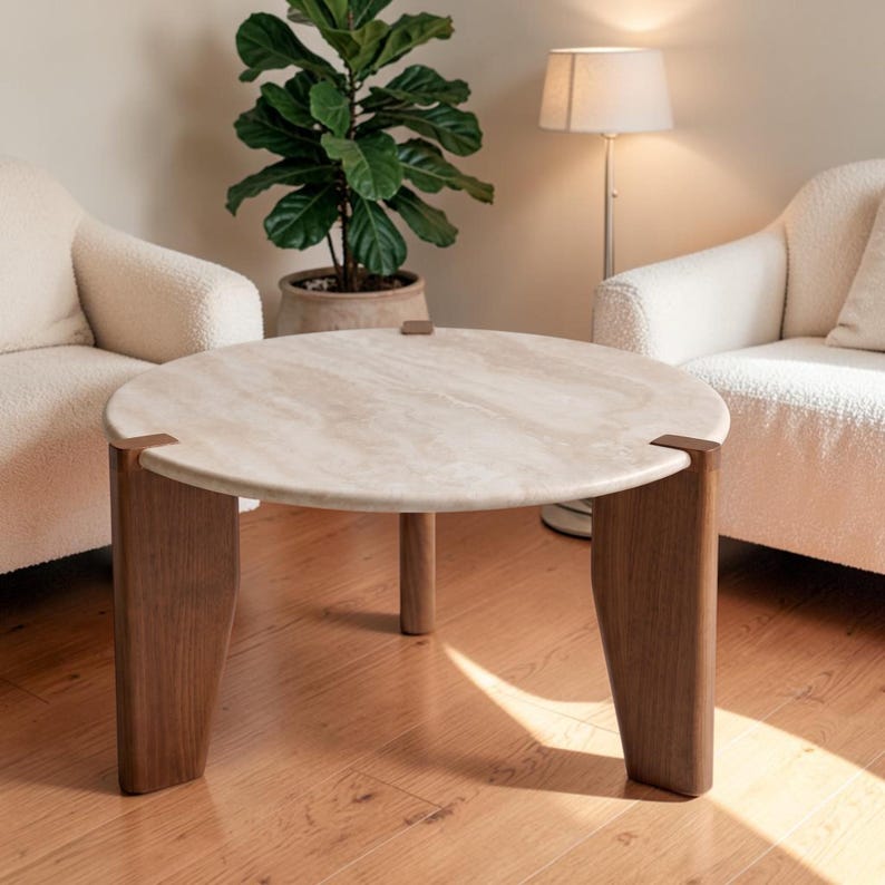 May include: A round coffee table featuring a beige, textured stone top with visible natural veining. The table is supported by three thick, dark brown wooden legs that connect to the stone surface with small, square metal brackets.
