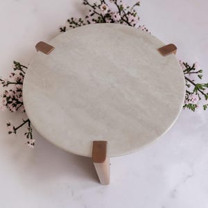 May include: A circular light gray stone pedestal stand featuring a smooth, polished surface. The stand is supported by three distinct, evenly spaced metallic copper-toned legs that grip the edge of the stone top, providing a stable base.