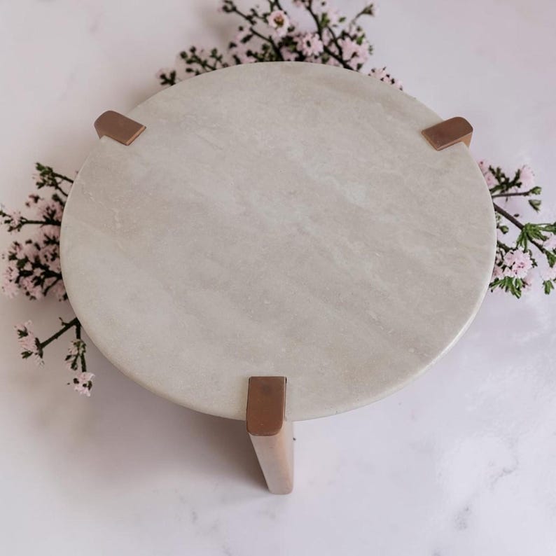 May include: A circular light gray stone pedestal stand featuring a smooth, polished surface. The stand is supported by three distinct, evenly spaced metallic copper-toned legs that grip the edge of the stone top, providing a stable base.
