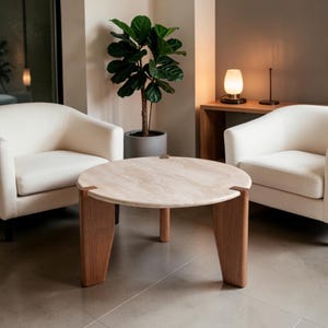May include: A round coffee table with a beige stone top and three thick wooden legs that extend through the surface. The table is positioned between two cream-colored upholstered armchairs, with a potted fiddle leaf fig plant nearby.