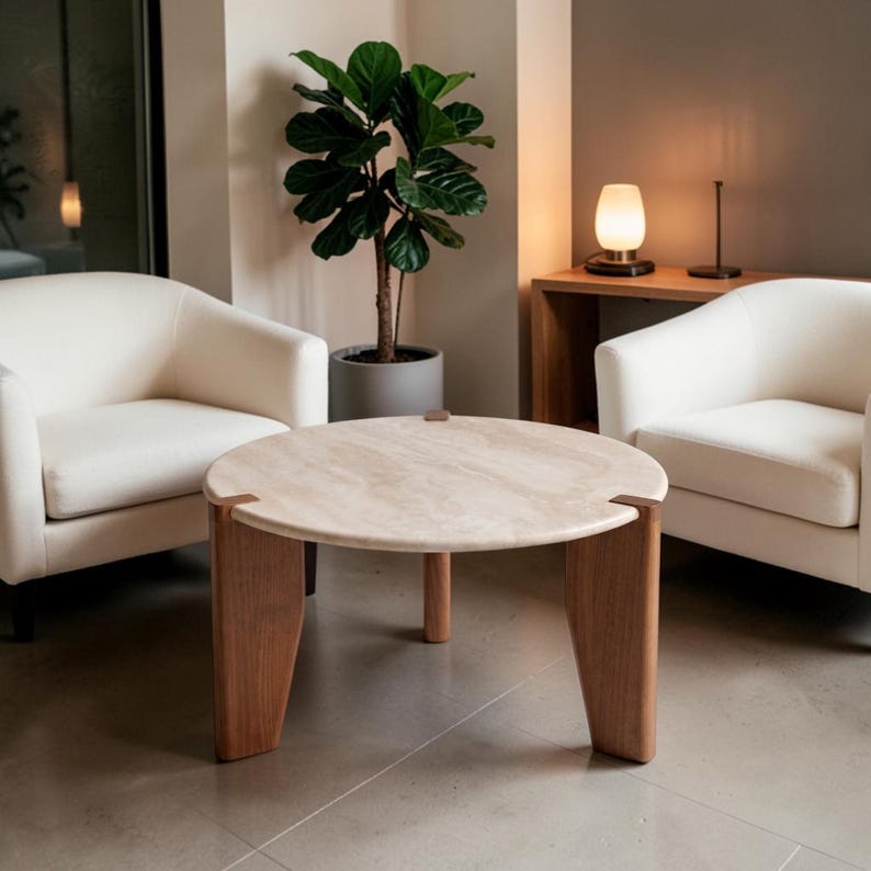 May include: A round coffee table with a beige stone top and three thick wooden legs that extend through the surface. The table is positioned between two cream-colored upholstered armchairs, with a potted fiddle leaf fig plant nearby.