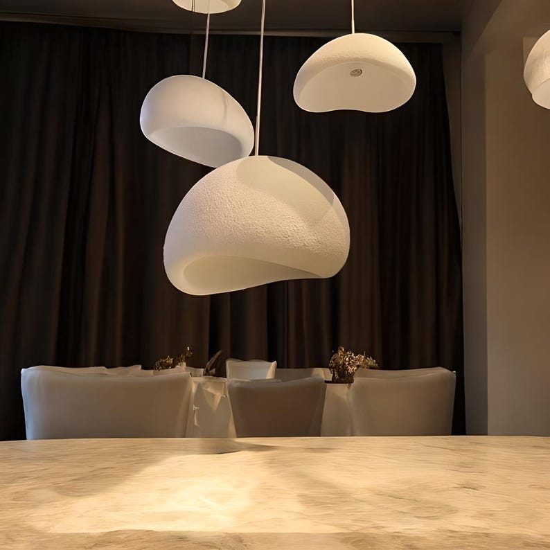 Pode incluir: Um conjunto de tr&ecirc;s luzes pendentes brancas de forma org&acirc;nica penduradas sobre uma mesa de jantar. As luzes s&atilde;o feitas de um material texturizado e t&ecirc;m um brilho suave e difuso.