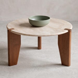 May include: A round coffee table with a light beige stone top and three dark brown wooden legs that slot into the edge of the surface. A matte sage green ceramic bowl sits in the center of the table, which has a smooth, polished finish.