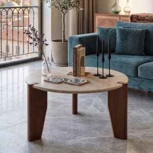May include: A round coffee table with a light beige stone top and three dark brown wooden legs. On the table are two square decorative tiles with floral patterns, a clear glass vase with dried flowers, two books, and three thin black candle holders.