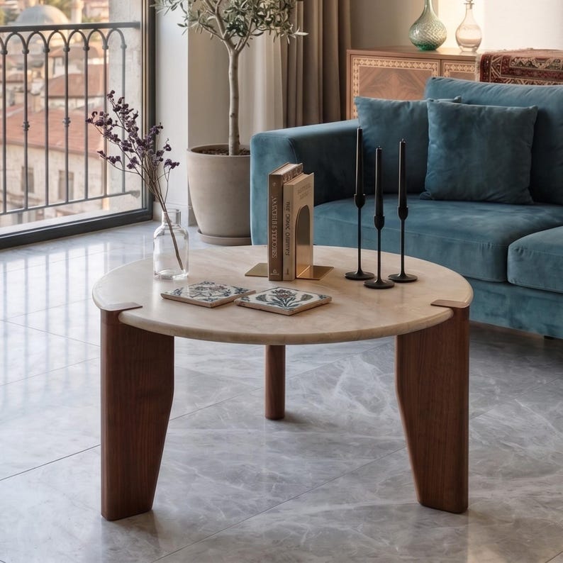 May include: A round coffee table with a light beige stone top and three dark brown wooden legs. On the table are two square decorative tiles with floral patterns, a clear glass vase with dried flowers, two books, and three thin black candle holders.