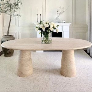 May include: A large oval travertine dining table with two pedestal bases. The table is set with a vase of white roses.