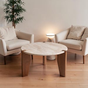 May include: A round coffee table with a light beige stone top and three dark brown wooden legs. The table is positioned between two cream-colored upholstered armchairs with matching throw pillows, creating a balanced seating arrangement.