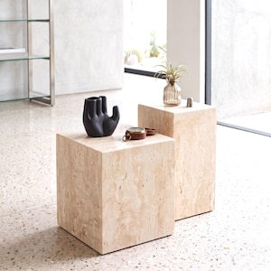 Puede incluir: Dos mesas cubo de travertino beige de diferentes alturas. La mesa más baja sostiene un jarrón negro mate con forma de mano. La mesa más alta sostiene un pequeño jarrón de cristal con una planta y un pequeño objeto metálico. Textura porosa.