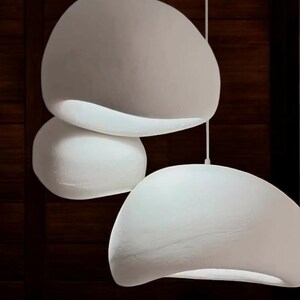 Pode incluir: Tr&ecirc;s luzes pendentes brancas de forma org&acirc;nica penduradas no teto. As luzes s&atilde;o suspensas por um acess&oacute;rio de teto branco. As luzes est&atilde;o em frente a uma parede de madeira escura.