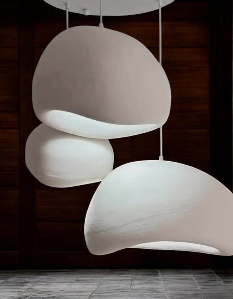 Pode incluir: Tr&ecirc;s luzes pendentes brancas de forma org&acirc;nica penduradas no teto. As luzes s&atilde;o suspensas por um acess&oacute;rio de teto branco. As luzes est&atilde;o em frente a uma parede de madeira escura.