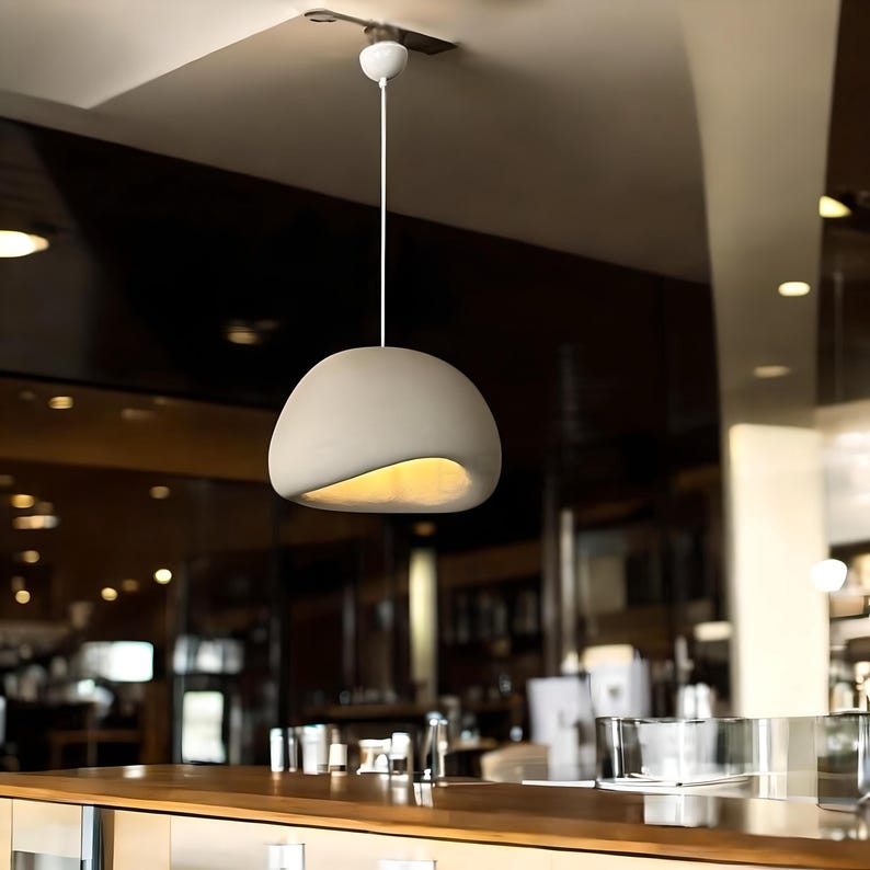 Pode incluir: Uma lumin&aacute;ria pendente moderna de concreto de cor branco-queimado pendura do teto em um ambiente comercial. A lumin&aacute;ria tem um formato org&acirc;nico &uacute;nico com uma abertura curva em forma de onda.