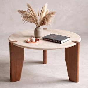 May include: A round coffee table with a light beige stone top and three thick, dark brown wooden legs. On the table sits a small gray vase filled with dried pampas grass, a small copper-colored tray with a white object, and a black book titled Sessiz Rüzgâr.