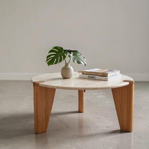 May include: A round coffee table with a light-colored stone top and three thick, angled wooden legs. On the table sits a small, round, light-colored vase containing a green monstera leaf and two stacked books titled Mid-Century Modern Design and EAMES.