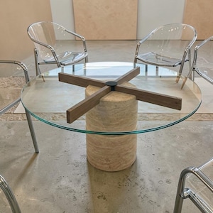 Op de afbeelding: Een moderne ronde glazen tafel met een lichtbruine houten basis en een kruisvormige steun. Heldere plastic stoelen met zilveren metalen poten omringen de tafel. De diameter van de tafel is ongeveer 122 cm.