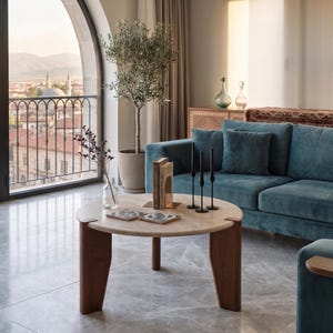 May include: A round coffee table with a light beige stone top and three dark brown wooden legs. On the table are two square patterned coasters, a clear glass vase with dried flowers, a book, and three tall, thin black metal candle holders.