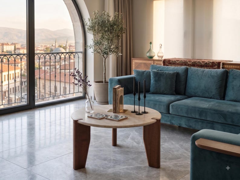 May include: A round coffee table with a light beige stone top and three dark brown wooden legs. On the table are two square patterned coasters, a clear glass vase with dried flowers, a book, and three tall, thin black metal candle holders.