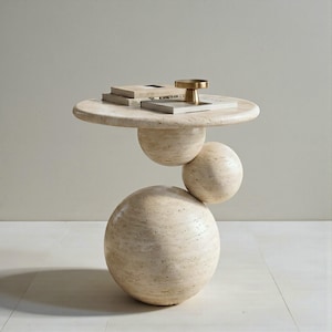May include: A beige travertine side table with a round top and three spherical supports. The table holds books and a small gold-coloured decorative object. The table's design is modern and sculptural, suitable for a living room or bedroom.