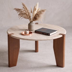May include: A round coffee table with a light beige stone top and three thick, dark brown wooden legs. On the table sits a small beige vase with dried pampas grass, a small copper-colored tray with a white object, and a black book titled 'Sesat Bangle'.