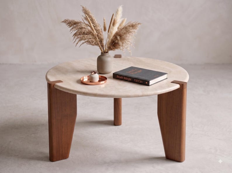 May include: A round coffee table with a light beige stone top and three thick, dark brown wooden legs. On the table sits a small beige vase with dried pampas grass, a small copper-colored tray with a white object, and a black book titled 'Sesat Bangle'.