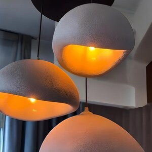 Pode incluir: Tr&ecirc;s luzes pendentes redondas, texturizadas e brancas com um suporte de teto preto. Cada luz tem um brilho amarelo quente.