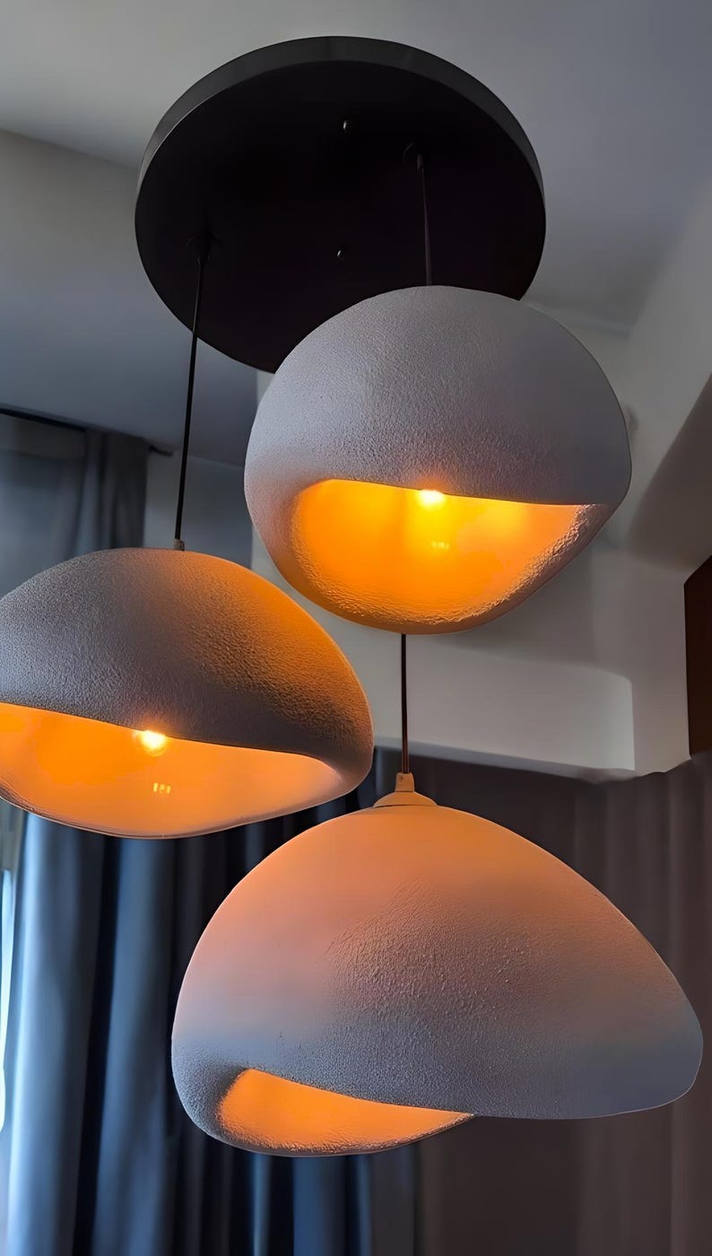 Pode incluir: Tr&ecirc;s luzes pendentes redondas, texturizadas e brancas com um suporte de teto preto. Cada luz tem um brilho amarelo quente.