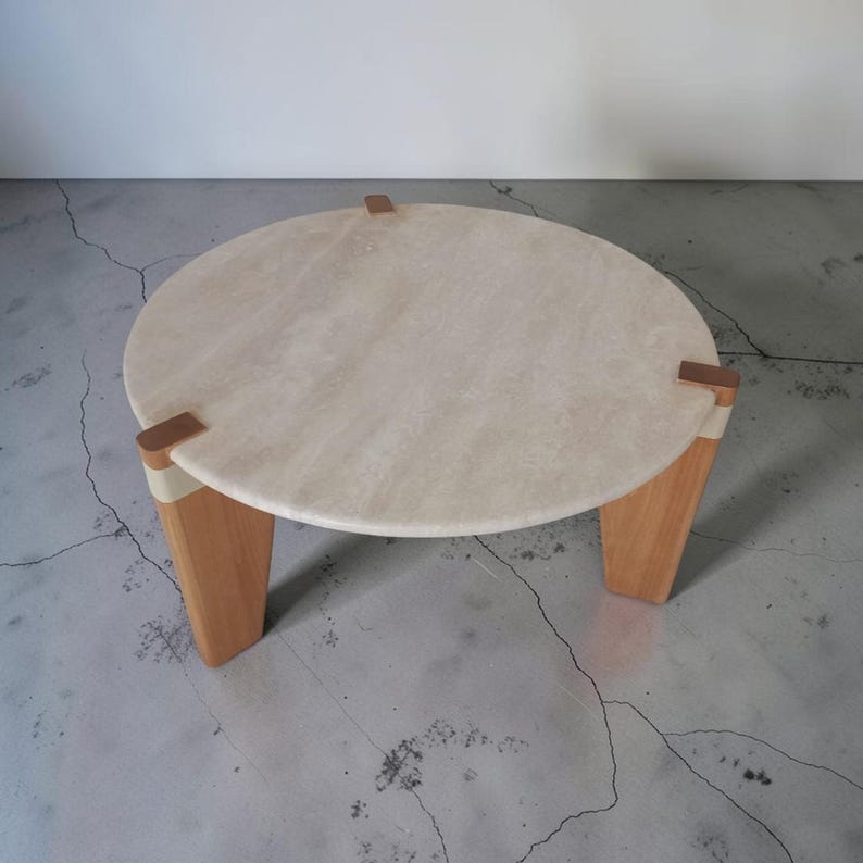 May include: A round coffee table featuring a beige travertine stone top with a textured surface. The table is supported by three light-toned wooden legs, each connected to the stone top with a small, rectangular metallic accent piece.