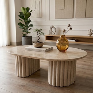 Könnte beinhalten: Ein ovaler, beigefarbener Couchtisch mit zylindrischem Sockel. Der Tisch hat eine hellfarbene Marmorplatte. Ein Bonsai-Baum, ein Buch mit dem Titel "Minimal Luxe" und eine bernsteinfarbene Glasvase stehen auf dem Tisch.