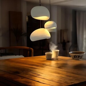 Pode incluir: Tr&ecirc;s luzes pendentes brancas, redondas, com forma de seixo, penduradas sobre uma mesa de madeira com duas x&iacute;caras de cer&acirc;mica branca e uma tigela na superf&iacute;cie.